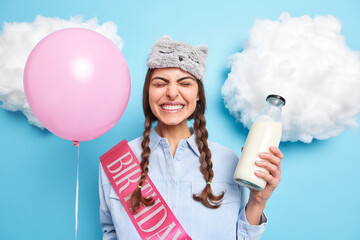 Positive teenage girl enjoys festive occasion grins at camera shows white teeth holds pink inflated balloon going to drink fresh milk dressed in domestic clothes isolated over blue background