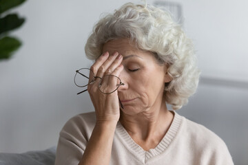 Tired senior lady holding glasses and rubbing face with closed eyes at home. Confused middle aged...