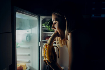 the girl opens and eats a banana next to the refrigerator. hunger concept. Looking for an overnight...