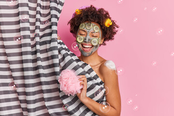 Positive Afro American woman sings song while showering applies cucumber facial mask to rejuvenate skin holds sponge enjoys body care procedures isolated on pink background soap bubbles flying around