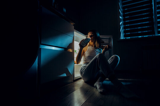 a hungry girl sits with her elbows on the open refrigerator and drinks milk. The image was shot in a low key to convey night time.