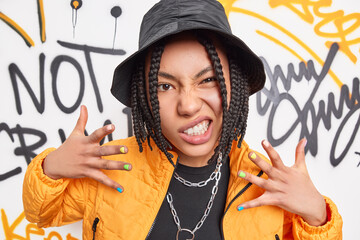 Rock style and urban lifestyle concept. Cheeky dark skinned woman smirks face raises hands enjoys rap music dressed in fashionable outfit poses against graffiti wall. Girl teenager with dreadlocks - obrazy, fototapety, plakaty
