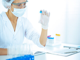 Professional female scientist in protective eyeglasses researching tube with reagents in laboratory toned in blue. Concepts of medicine and science researching