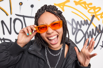 Trendy cheerful dark skinned hipster girl wears orange sunglasses black jacket raises hand exclaims loudly has cheeky expression has own fashion style poses against creative painted street wall - obrazy, fototapety, plakaty