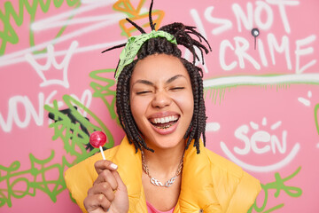 Trendy cheerful hipster girl smiles broadly holds lollipop has fun with teenagers of same age wears yellow vest poses against colorful graffiti wall. Gen z generation. Youth lifestyle concept - obrazy, fototapety, plakaty