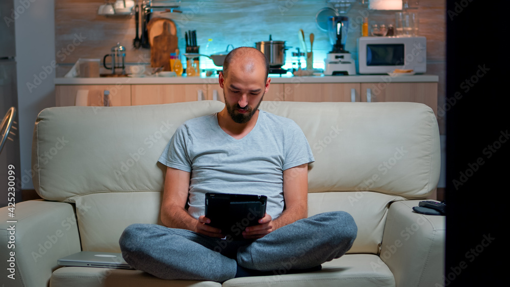 Aufkleber tired man sitting alone on couch while browsing on internet using tablet computer with modern techno
