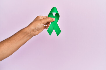 Hand of young hispanic man holding green ribbon over isolated pink background.