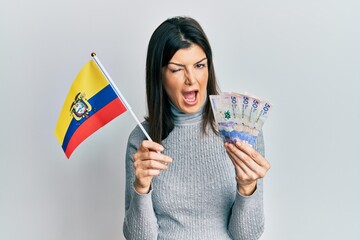 Young hispanic woman holding colombia flag and colombian pesos banknotes winking looking at the...