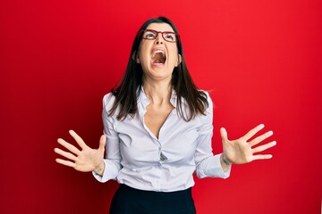 Young hispanic woman wearing business shirt and glasses crazy and mad shouting and yelling with aggressive expression and arms raised. frustration concept.