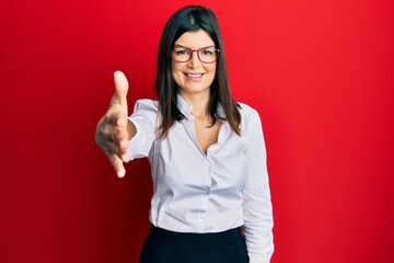 Young hispanic woman wearing business shirt and glasses smiling friendly offering handshake as greeting and welcoming. successful business.