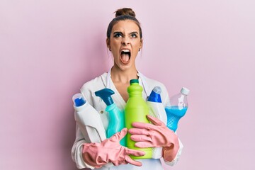 Beautiful brunette young woman wearing cleaner apron holding cleaning products angry and mad...