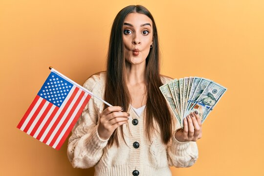 Beautiful brunette young woman holding united states flag and dollars making fish face with mouth and squinting eyes, crazy and comical.