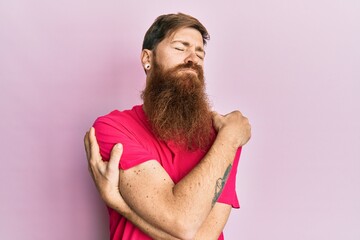 Redhead man with long beard wearing casual pink t shirt hugging oneself happy and positive, smiling confident. self love and self care