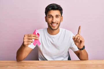 Young handsome man holding pink cancer ribbon surprised with an idea or question pointing finger...