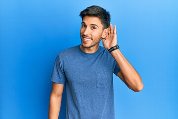 Young handsome man wearing casual tshirt over blue background smiling with hand over ear listening and hearing to rumor or gossip. deafness concept.
