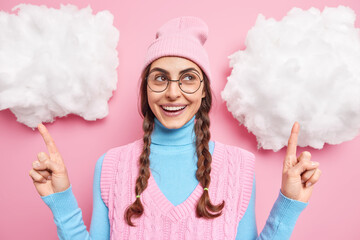 Positive beautiful European girl with pigtails points above on white clouds demonstrates something wears hat turtleneck round spectacles and vest isolated over pink background shows something good.
