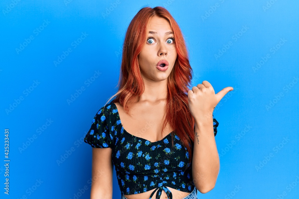 Poster young beautiful redhead woman wearing casual clothes over blue background surprised pointing with ha