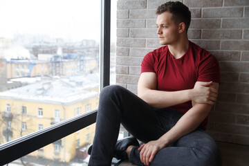 A nice guy in burgundy casual clothes sits against the background of a panoramic window on a winter day.