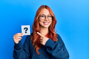 Young beautiful redhead woman holding question mark smiling happy pointing with hand and finger