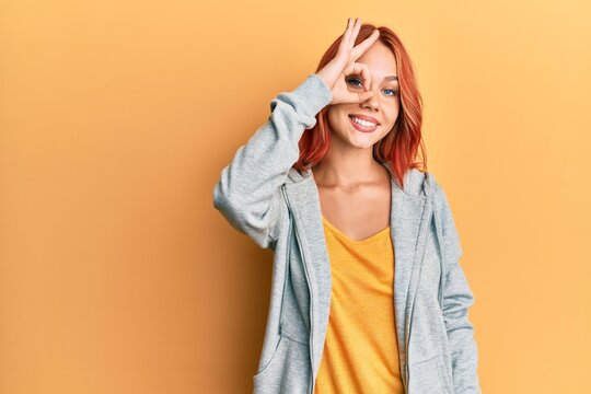 Young beautiful redhead woman wearing casual sporty sweatshirt smiling happy doing ok sign with hand on eye looking through fingers