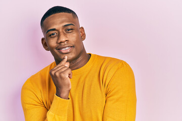 Young black man wearing casual yellow sweater smiling looking confident at the camera with crossed arms and hand on chin. thinking positive.