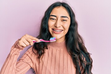 Hispanic teenager girl with dental braces holding mouthwash and toothbrush for fresh breath looking...
