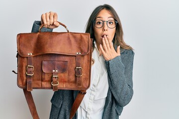 Young brunette woman holding leather bag covering mouth with hand, shocked and afraid for mistake....