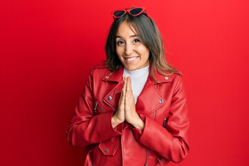 Young brunette woman wearing red leather jacket praying with hands together asking for forgiveness smiling confident.