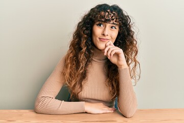 Young hispanic girl wearing casual clothes sitting on the table serious face thinking about...