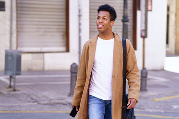 Young black man walking down the street carrying a briefcase and a smartphone in his hand.