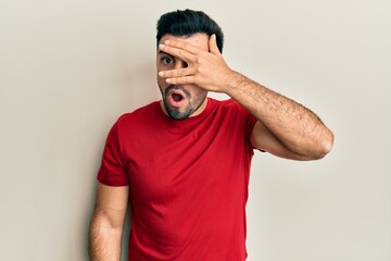 Young hispanic man wearing casual clothes peeking in shock covering face and eyes with hand,...