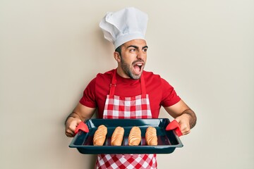 Young hispanic man wearing baker uniform holding homemade bread angry and mad screaming frustrated and furious, shouting with anger. rage and aggressive concept.
