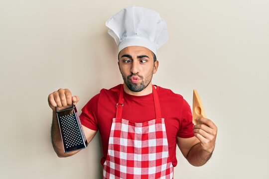 Young hispanic man wearing professional cook uniform holding grater and cheese making fish face with mouth and squinting eyes, crazy and comical.