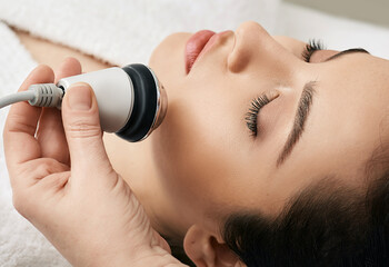 Brunette woman receiving radiofrequency lifting procedure for her face skin rejuvenation at...