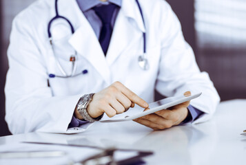 Unknown male doctor sitting and working with tablet computer in clinic at his working place, close-up. Young physician at work. Perfect medical service, medicine concept