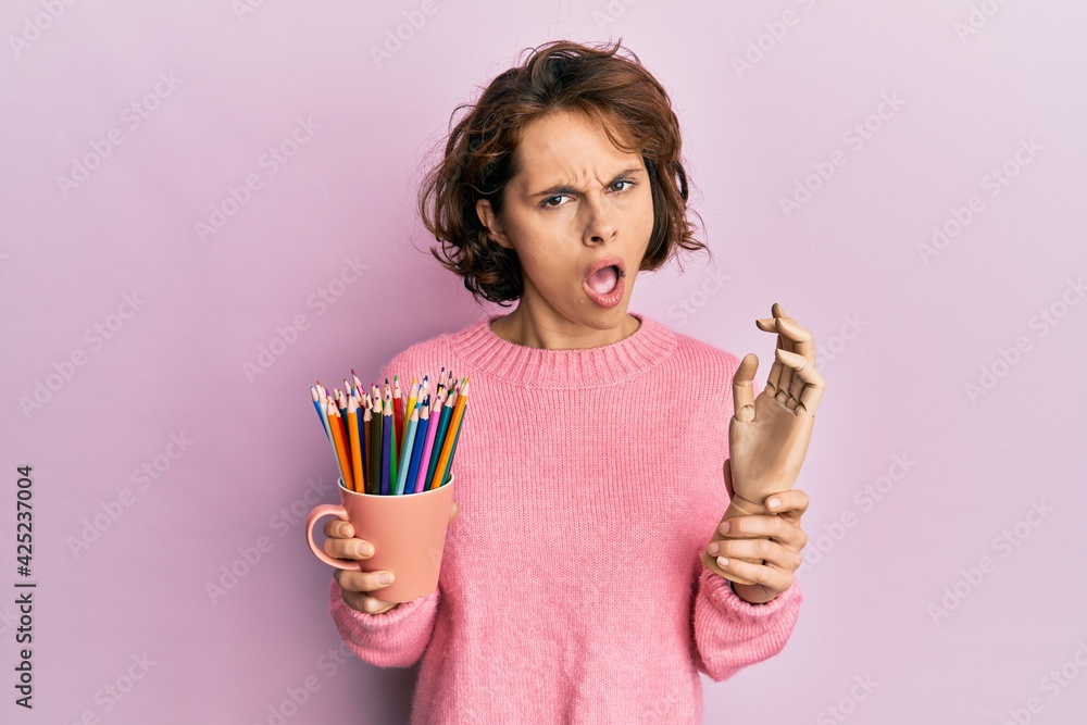 Poster Young brunette woman holding small wooden manikin hand and pencils in shock face, looking skeptical and sarcastic, surprised with open mouth