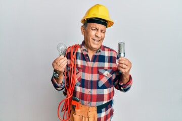 Senior hispanic man wearing handyman uniform holding led lightbulb and incandescent bulb winking...
