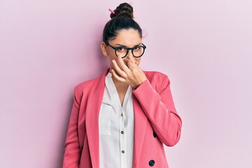 Beautiful middle eastern woman wearing business jacket and glasses smelling something stinky and disgusting, intolerable smell, holding breath with fingers on nose. bad smell