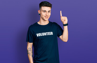 Fototapeta premium Young caucasian boy with ears dilation wearing volunteer t shirt with a big smile on face, pointing with hand finger to the side looking at the camera.
