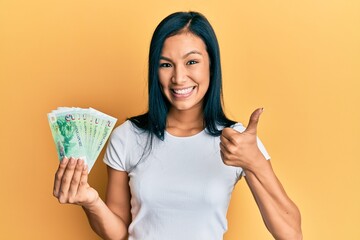 Beautiful hispanic woman holding 5 singapore dollars banknotes smiling happy and positive, thumb up doing excellent and approval sign