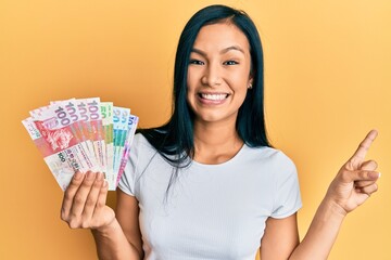 Beautiful hispanic woman holding hong kong dollars banknotes smiling happy pointing with hand and finger to the side