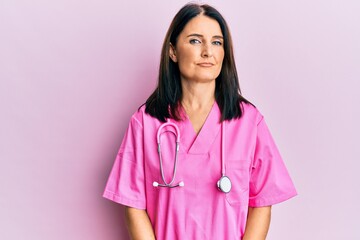 Middle age brunette woman wearing doctor uniform and stethoscope relaxed with serious expression on...