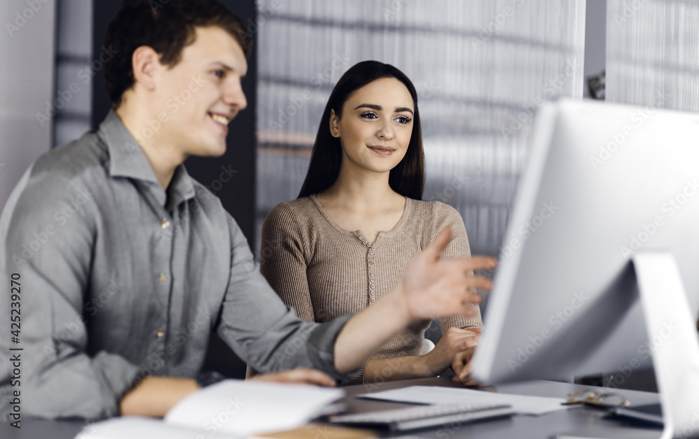 Wall mural Friendly young businessman and programmer in a green shirt is working on computer, while sitting together at the desk with a female colleague in a modern office. Focus on woman. Concept of successful