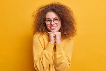 Smiling beautiful teenage girl has natural bushy curly hair smiles toothily keeps hands under chin wears spectacles casual hoodie in one tone with background being pleased by hearing good news