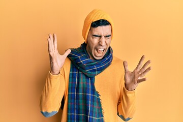 Handsome hispanic man wearing wool hat and winter scarf celebrating mad and crazy for success with arms raised and closed eyes screaming excited. winner concept