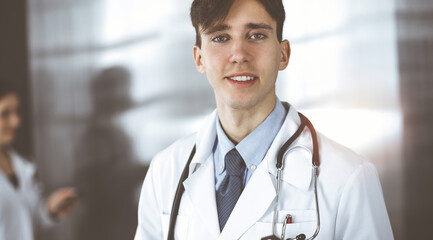 Friendly young male doctor standing with clipboard in modern clinic. Female colleague is busy with...