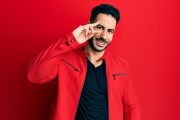 Young hispanic man wearing red leather jacket doing peace symbol with fingers over face, smiling cheerful showing victory