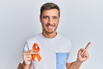 Handsome caucasian man holding awareness orange ribbon smiling happy pointing with hand and finger...