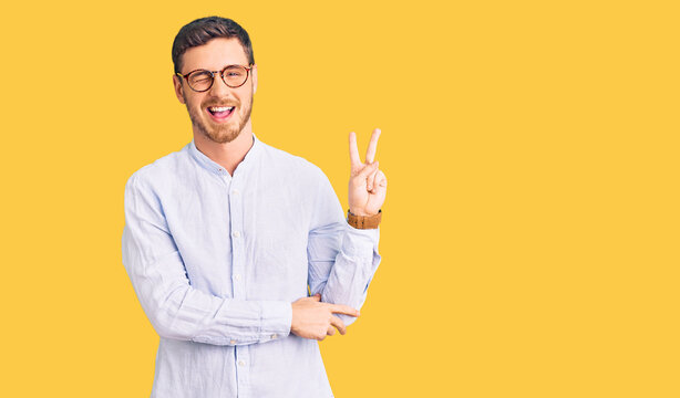 Handsome young man with bear wearing elegant business shirt and glasses smiling with happy face winking at the camera doing victory sign with fingers. number two.