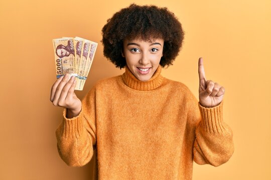 Young hispanic girl holding 5000 hungarian forint banknotes smiling with an idea or question pointing finger with happy face, number one